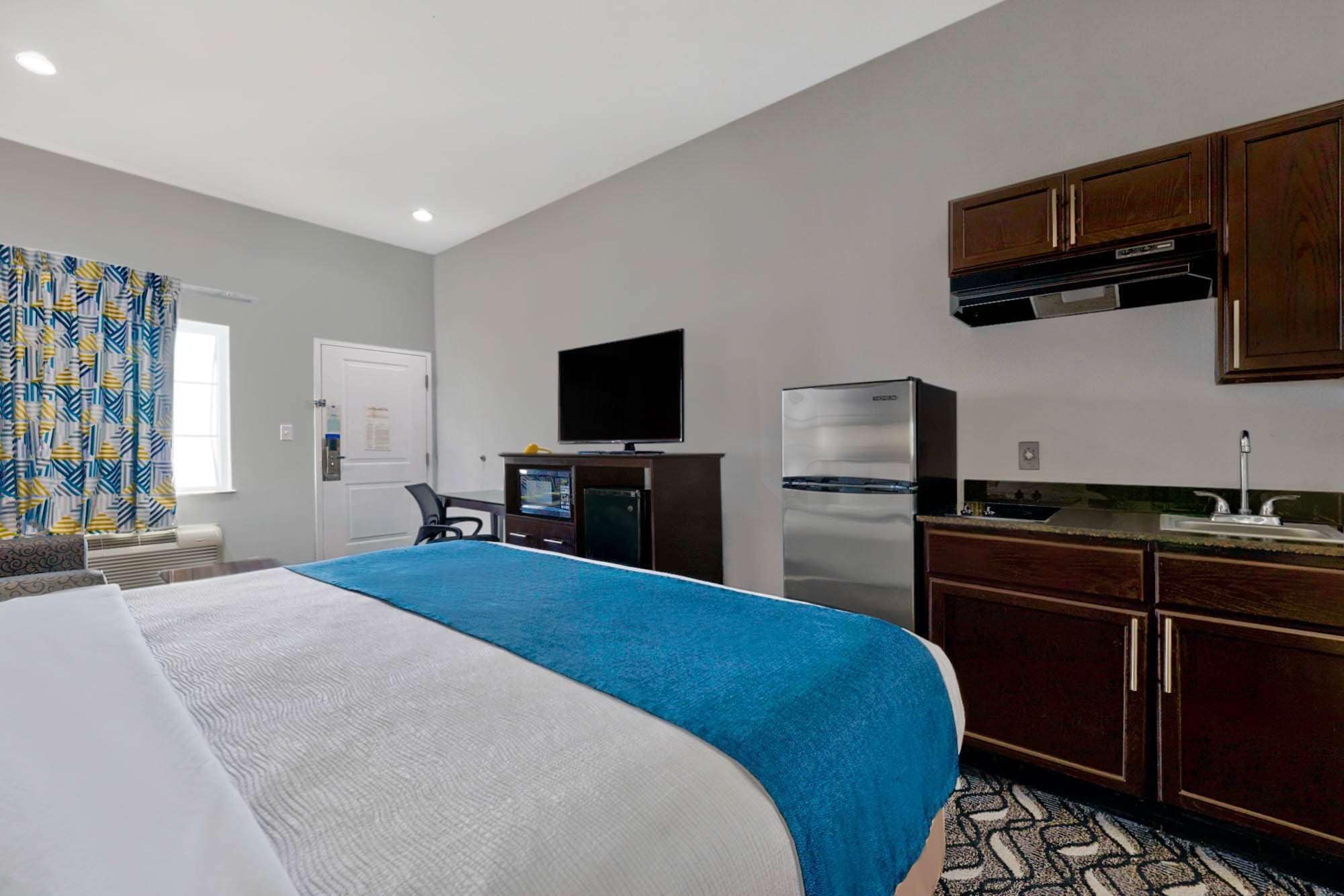 A hotel room with a bed, a desk with a computer, a TV, a refrigerator, and a kitchenette.