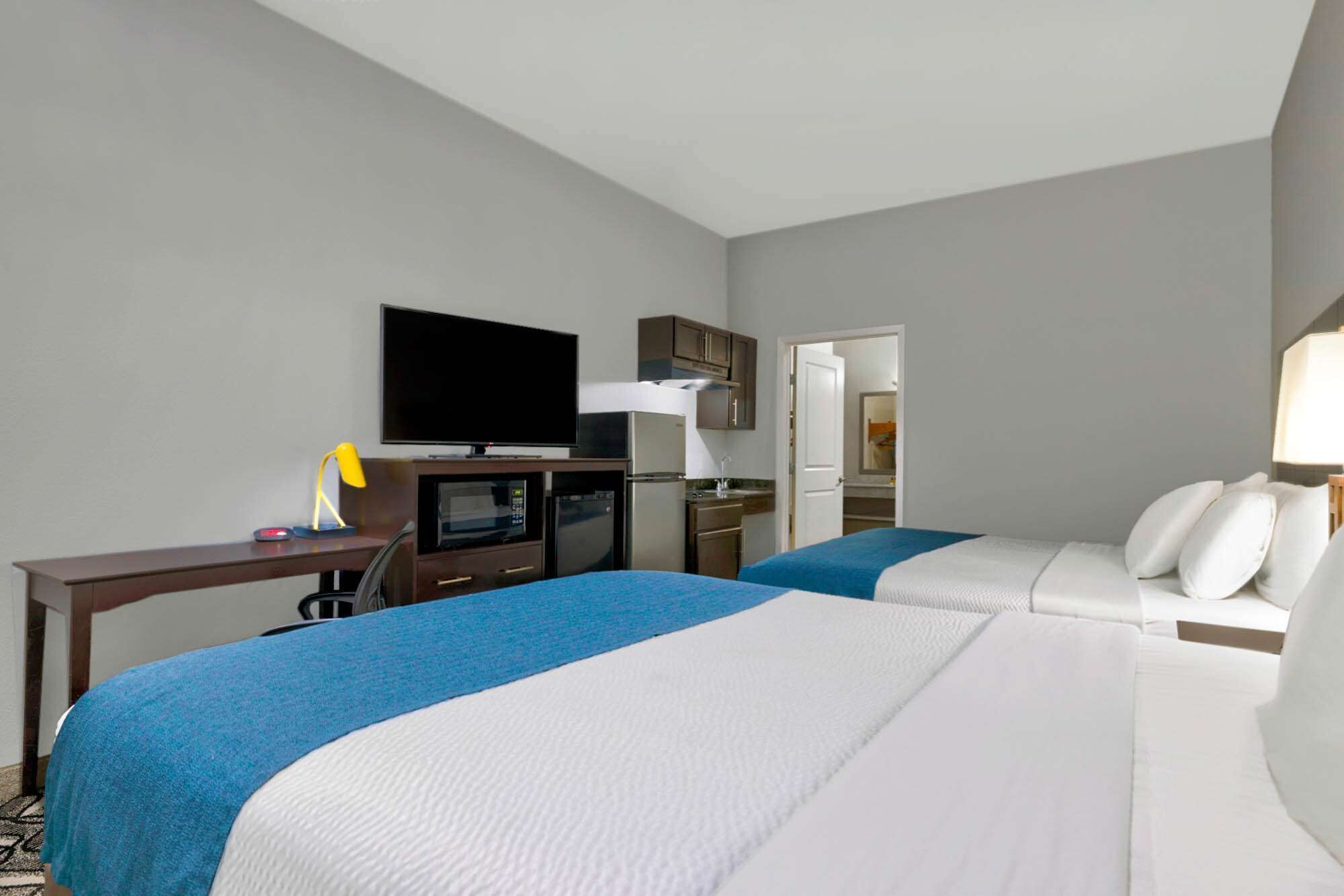 A hotel room with two beds, a TV, a desk, and a kitchenette.
