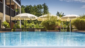 A swimming pool with umbrellas, surrounded by greenery and a building. -  feel good Resort Wörthersee (Krumpendorf am Woerther See)