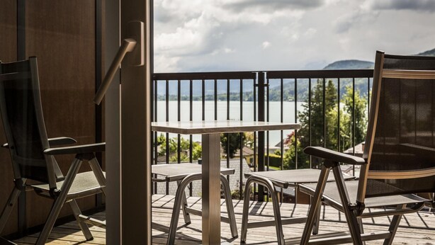 A balcony with a table and chairs, overlooking a body of water and greenery. -  feel good Resort Wörthersee (Krumpendorf am Woerther See)