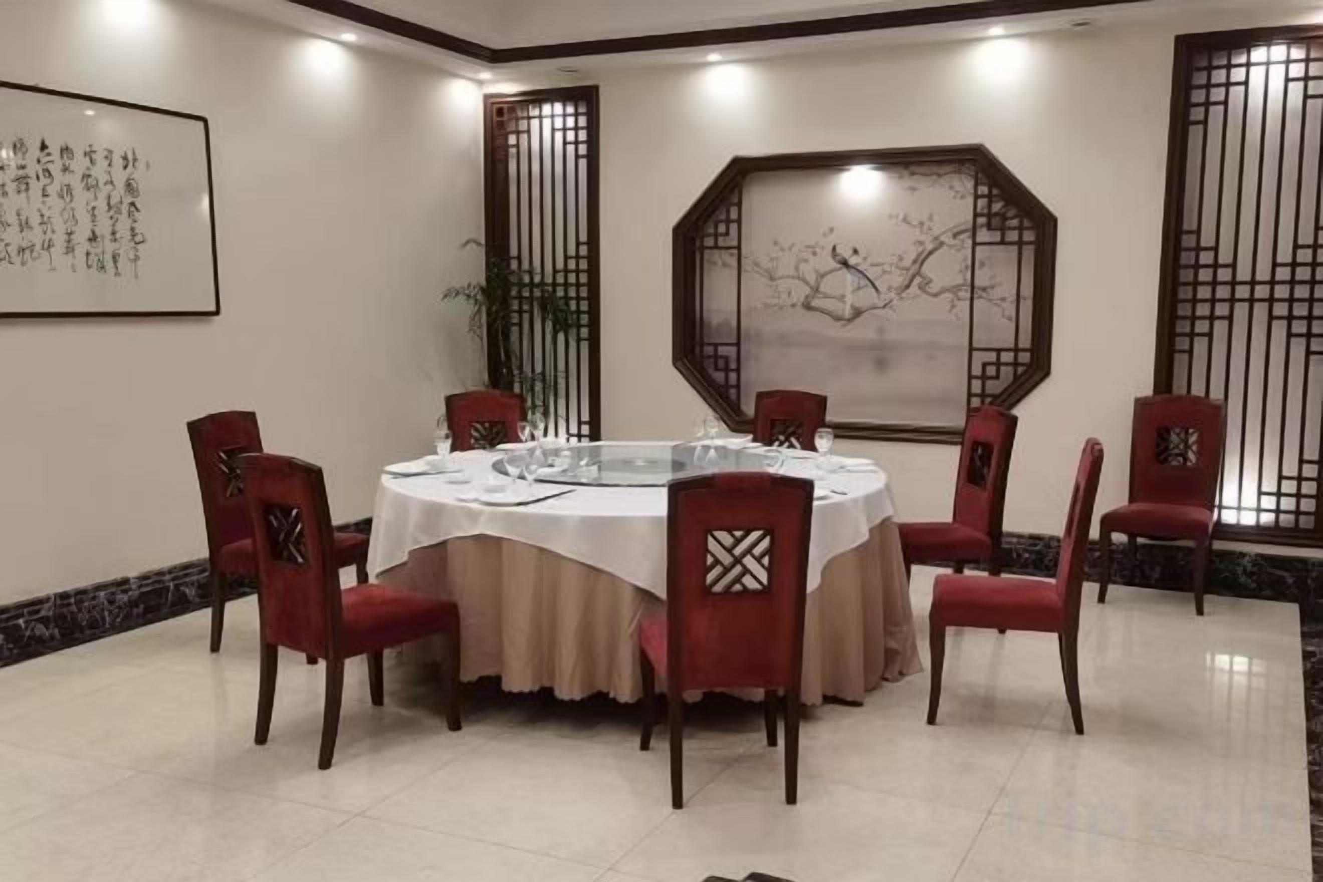 A round dining table with a white tablecloth, surrounded by six red chairs with patterned backrests, in a room with traditional decor and artwork on the walls.