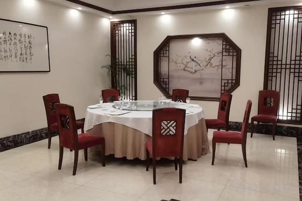 A round dining table with a white tablecloth, surrounded by six red chairs with patterned backrests, in a room with traditional decor and artwork on the walls. - Sihai Grand Hotel (Anshan)
