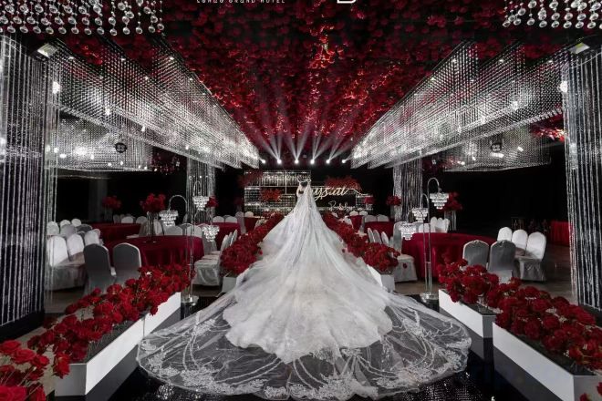 A wedding venue with a central aisle leading to a stage, surrounded by red floral arrangements and mirrored walls.