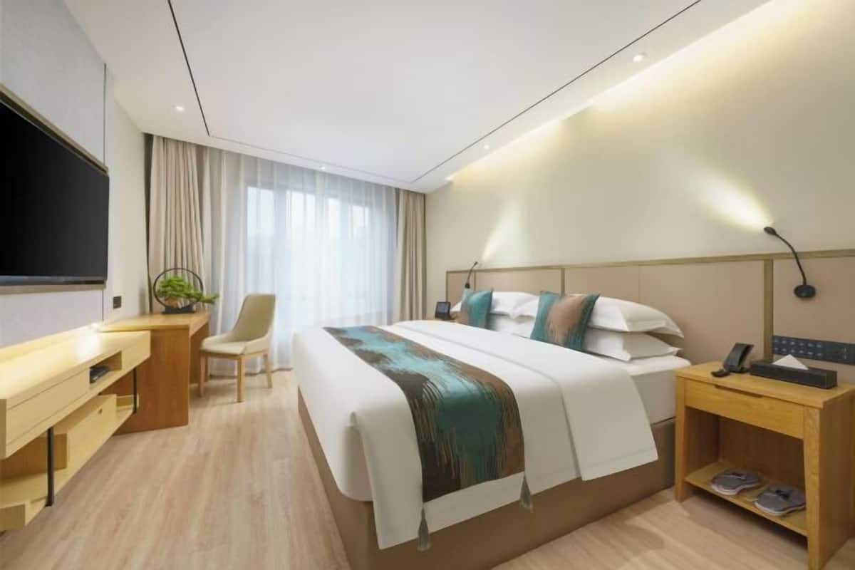 A modern hotel room with a bed, a desk, a chair, a TV, and a bedside table with a lamp.