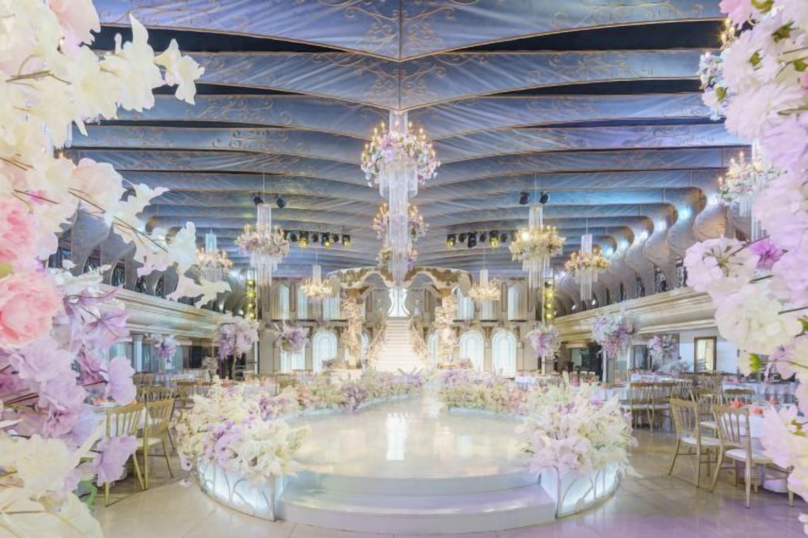 A grand hall with a central chandelier, ornate ceiling, and floral arrangements.