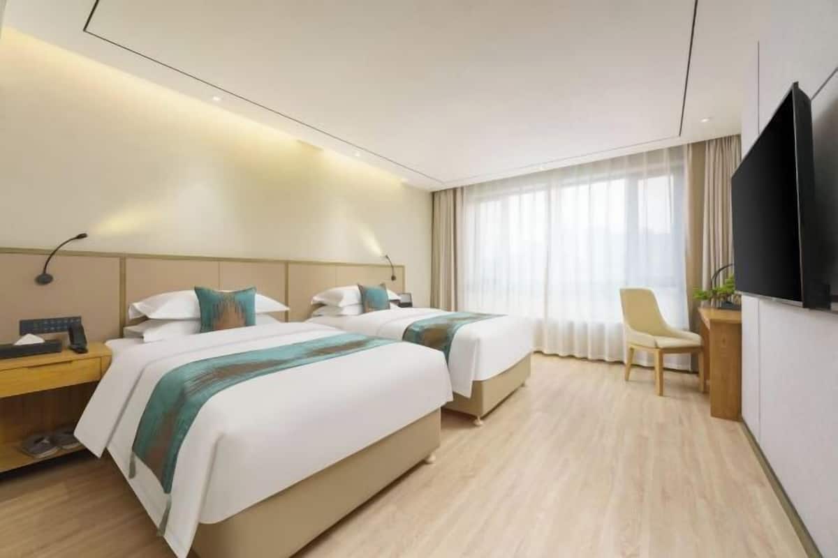 A hotel room with two beds, a desk, a chair, a TV, and a large window with curtains.