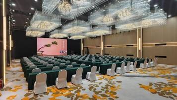 Un salón de conferencias con filas de mesas y sillas, un escenario con un telón de fondo rosa y una lámpara de araña grande.