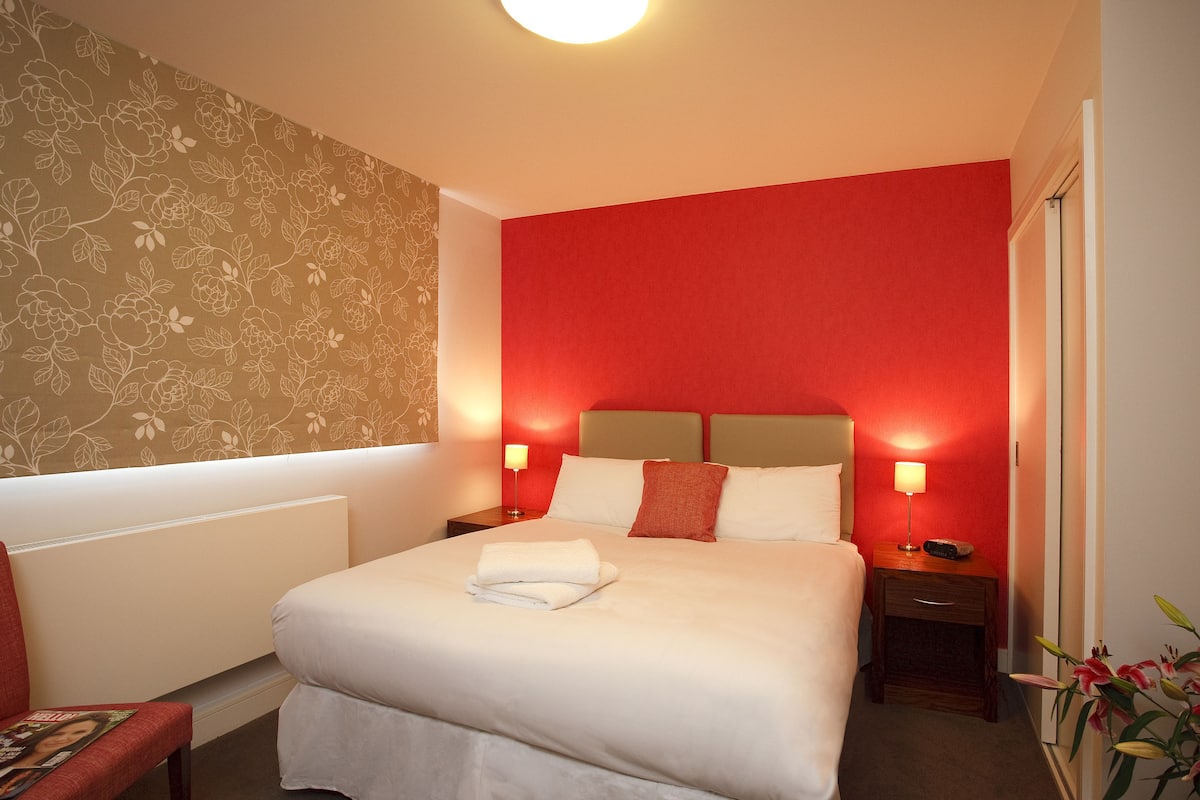 Ein Hotelzimmer mit einem großen Bett, zwei Nachttischen mit Lampen, einer roten Akzentwand und floralem Wandpapier auf der linken Seite.