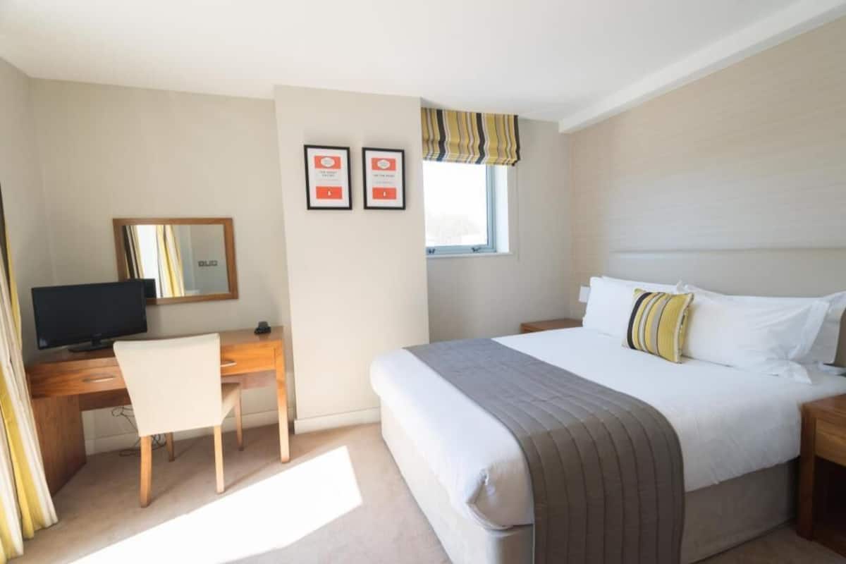 Ein Hotelzimmer mit einem Bett, einem Schreibtisch mit Computer, einem Stuhl, einem Fenster mit Vorhängen und zwei gerahmten Zertifikaten an der Wand.