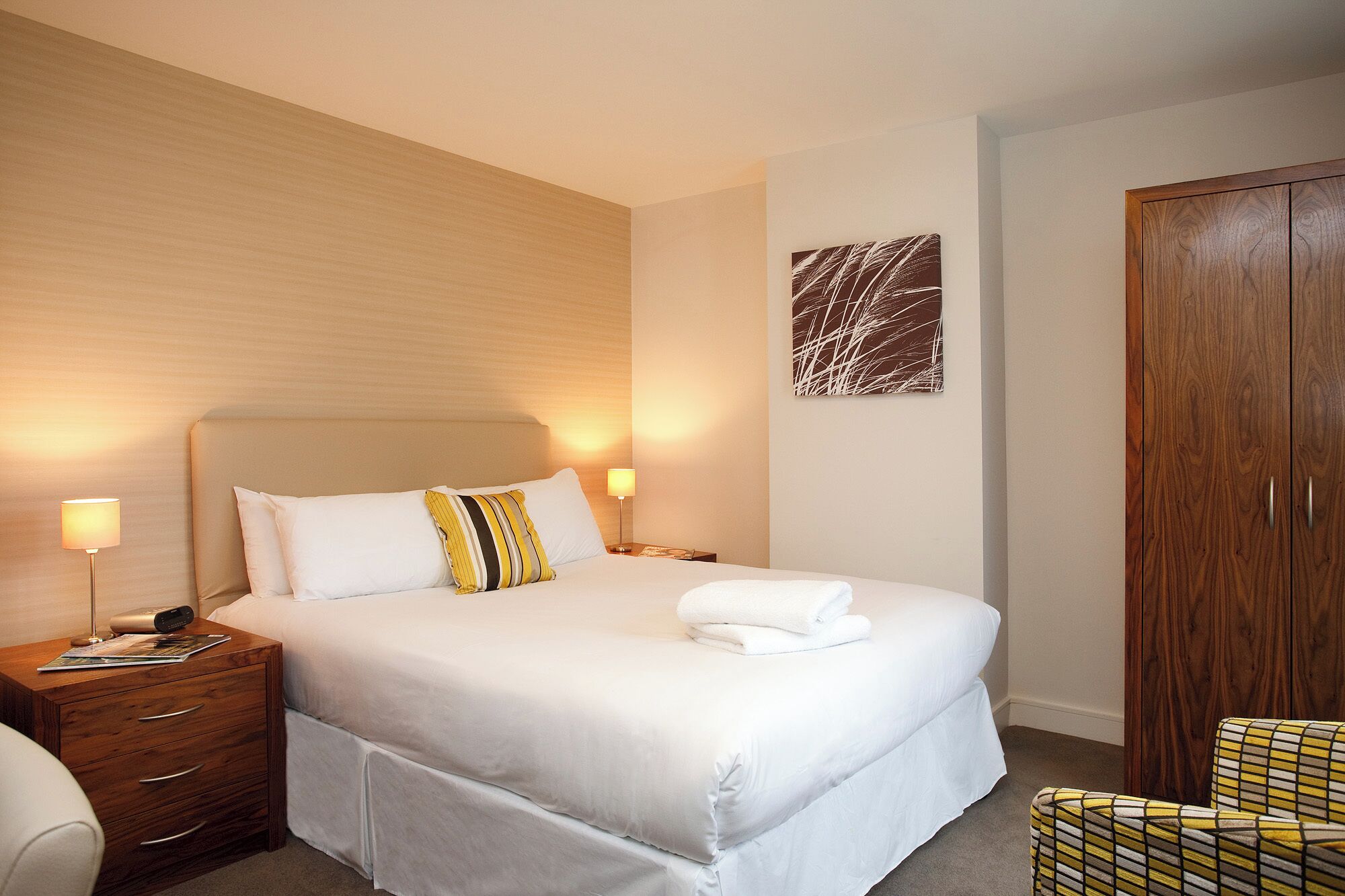 Quarto de hotel com uma cama grande, duas mesas de cabeceira, um guarda-roupa de madeira e uma obra de arte abstrata na parede.