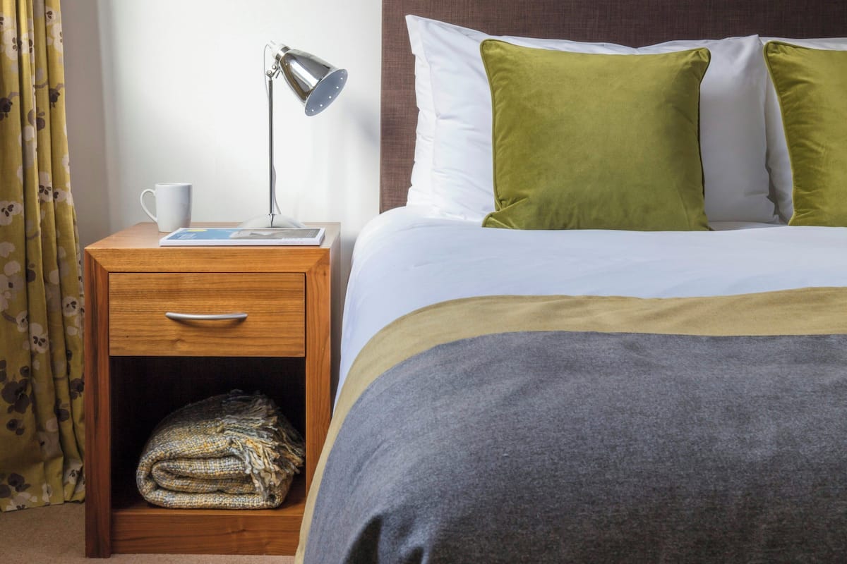 Ein Hotelzimmer mit Bett, Nachttisch, Lampe, Tasse und gefalteter Decke.