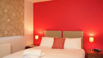 Une chambre d’hôtel avec un mur rouge, un lit recouvert de draps blancs, deux lampes de chevet et une table de chevet en bois sur laquelle se trouve un appareil Sony.