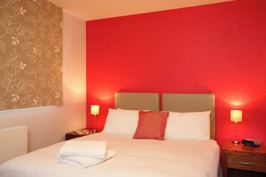 A hotel room with a red wall, a bed with white linens, two bedside lamps, and a wooden nightstand with a Sony device.