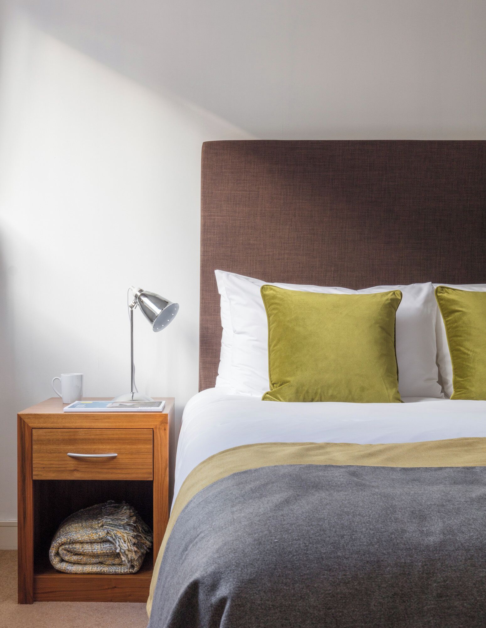 A neatly made bed with a grey blanket, a wooden nightstand with a lamp, a white mug, and a folded blanket in a drawer.