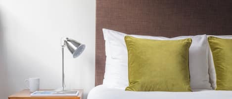 A neatly made bed with a grey blanket, a wooden nightstand with a lamp, a white mug, and a folded blanket in a drawer.
