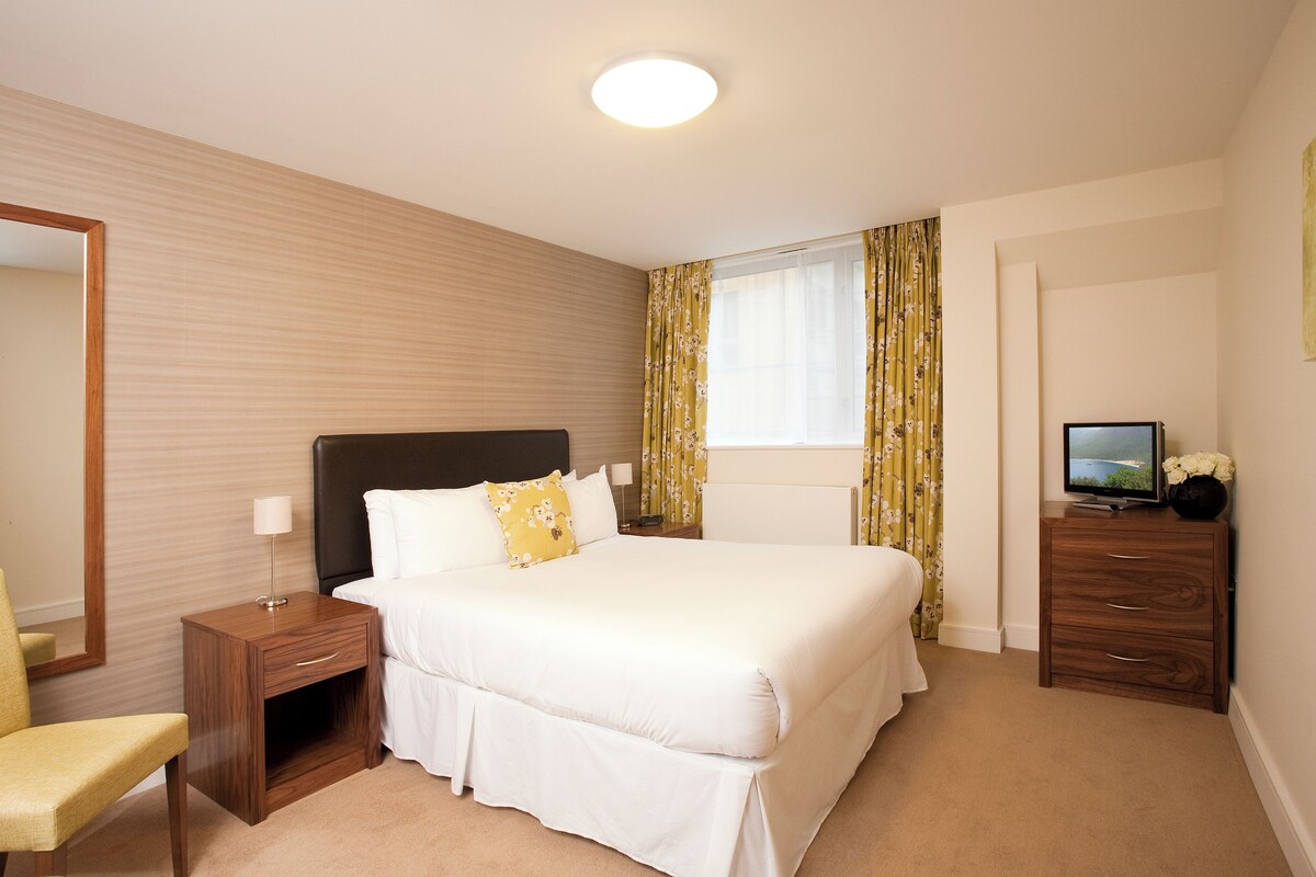 A hotel room with a large bed, a wooden nightstand, a chair, a television on a dresser, and a window with curtains.