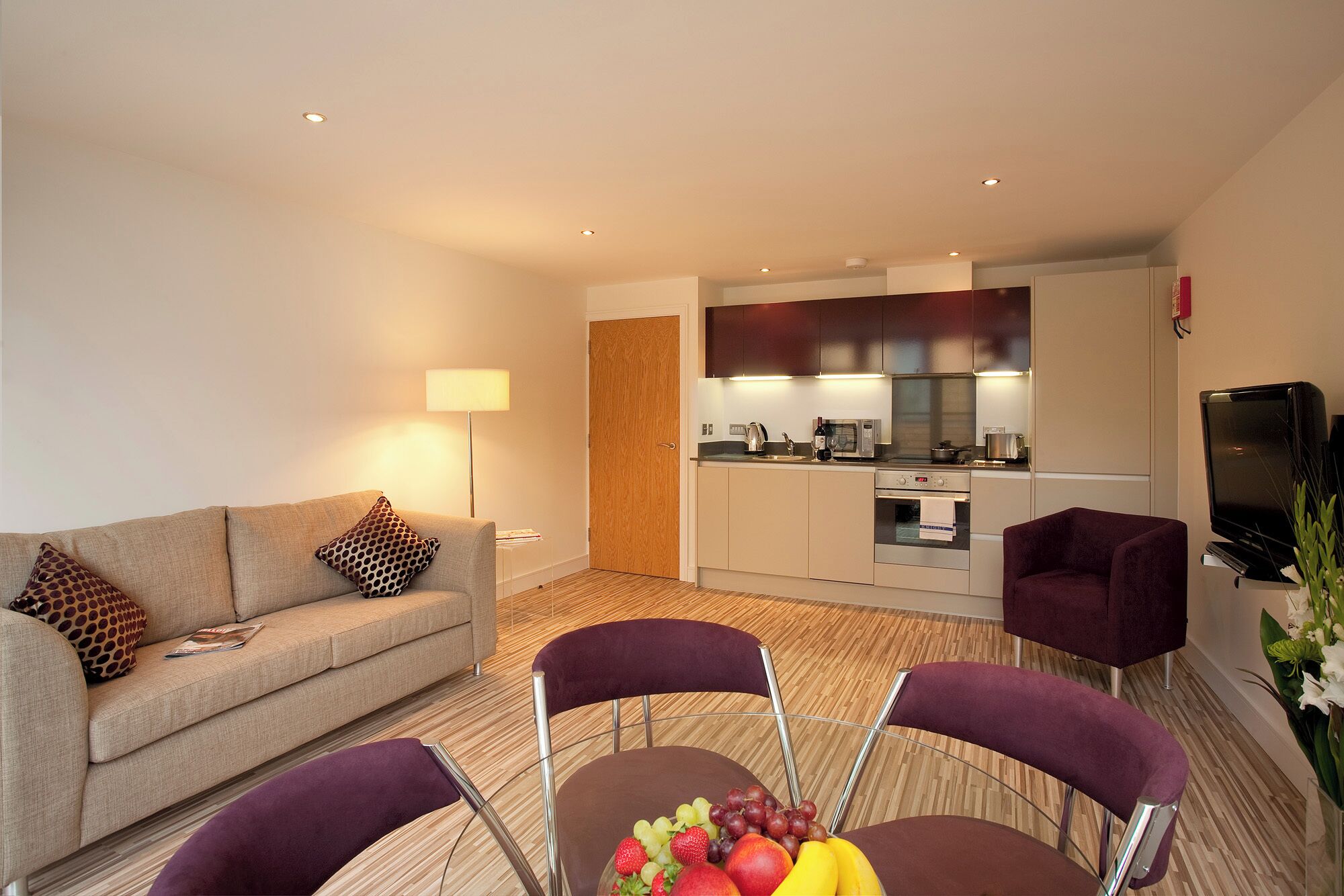 A modern living room with a sofa, two purple chairs, a dining table with fruit, and a kitchen area with appliances.
