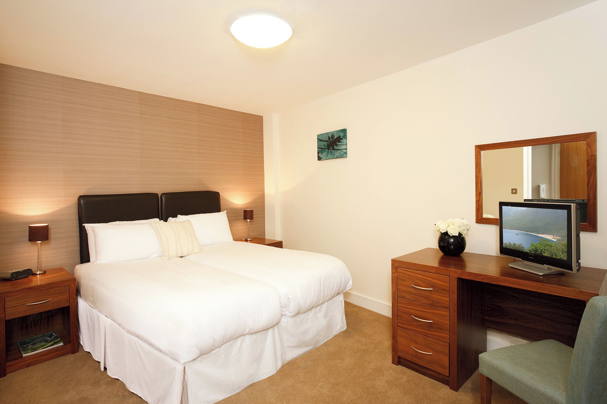 A hotel room with a bed, bedside tables, a desk with a computer, a chair, and a large mirror.