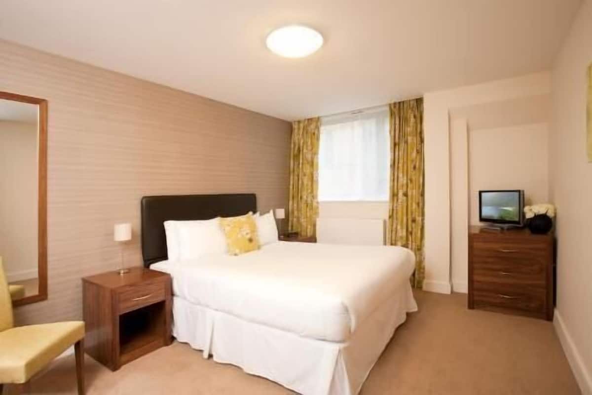 Ein Hotelzimmer mit Bett, Nachttisch, Sessel, Kommode, Fernseher und einem Fenster mit Vorhängen.