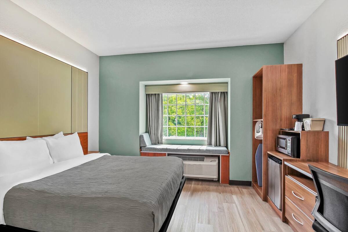 A hotel room with a bed, a desk with a chair, a TV, a microwave, and a window with curtains.