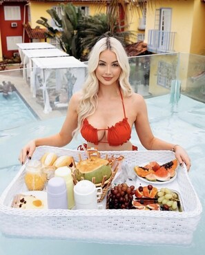 Une femme vêtue d’un bikini rouge, tenant un plateau sur lequel se trouvent divers aliments et une boisson, se trouve au bord d’une piscine.