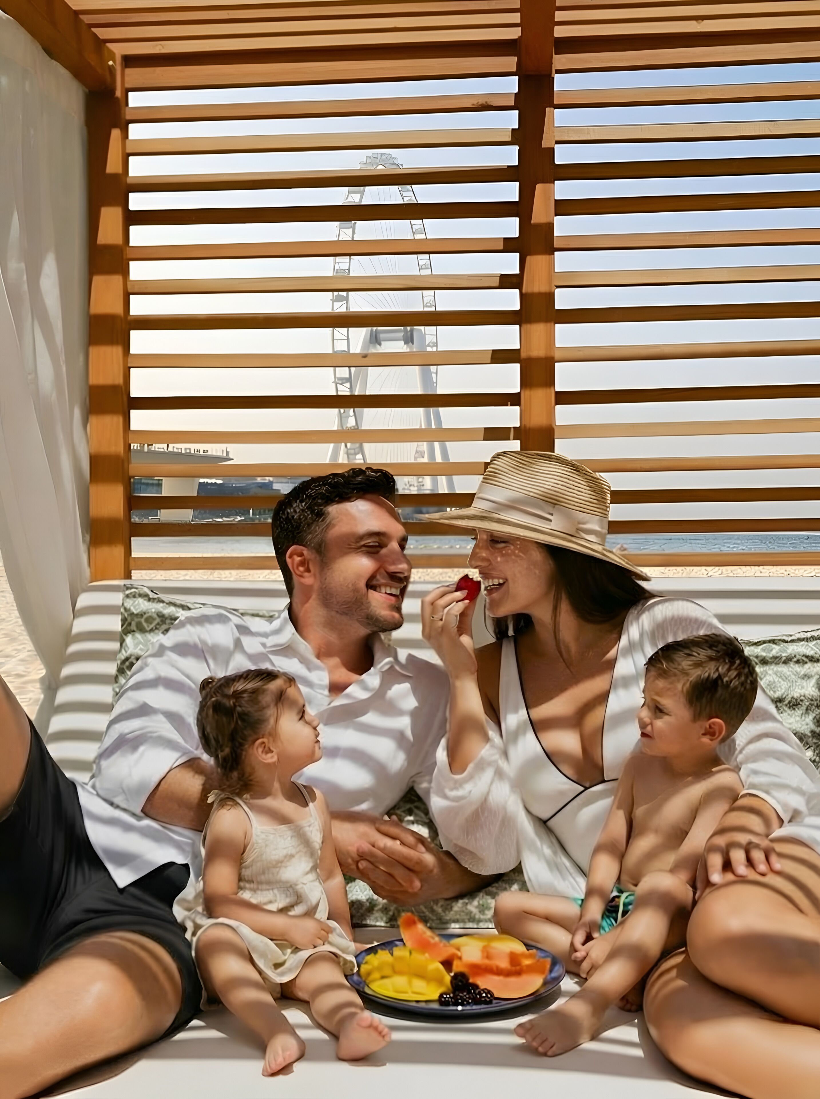 Une famille est assise sur une chaise longue blanche, avec une assiette de fruits, sous une pergola en bois dont les lattes sont disposées horizontalement.