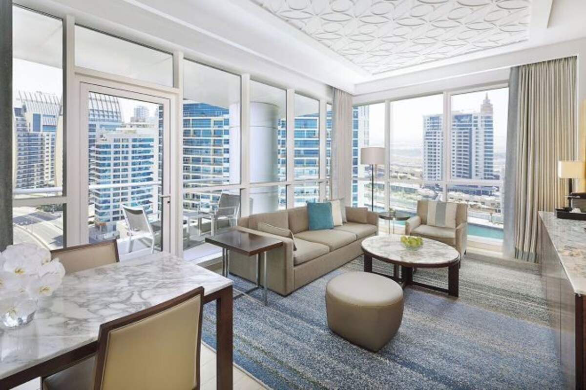 A spacious hotel room with a large balcony, a seating area with a sofa and armchairs, a dining table, and a view of the cityscape.