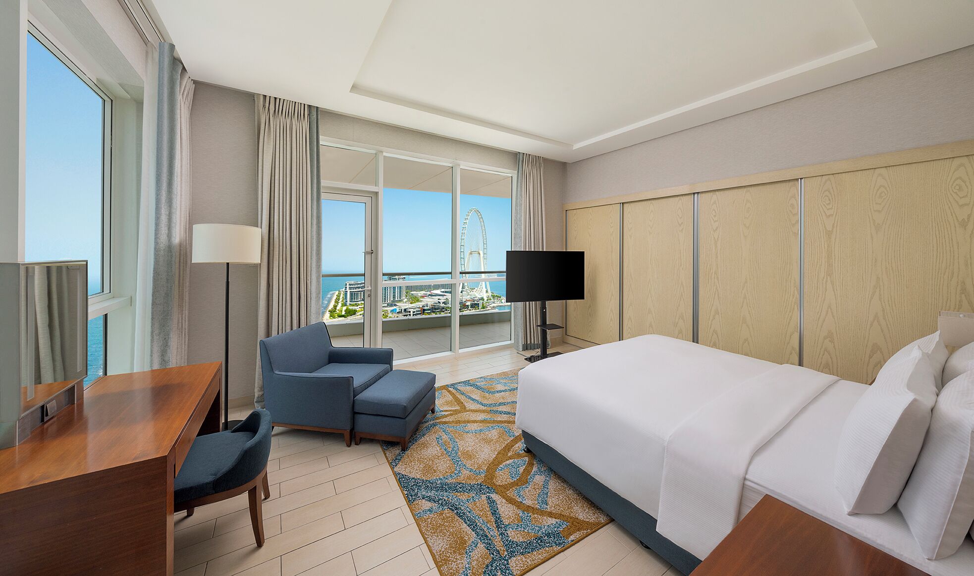 A hotel room with a bed, a desk, a chair, a TV, and a balcony with a view of a Ferris wheel and buildings.