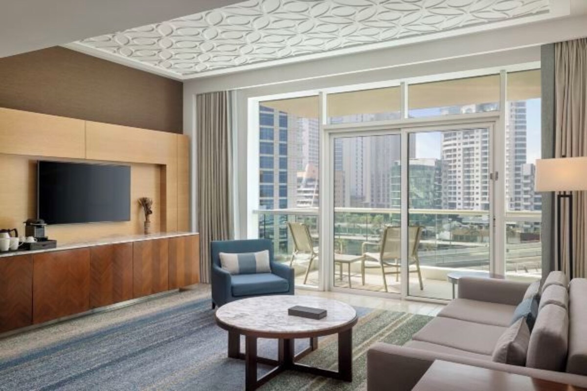 Une chambre d’hôtel moderne dotée d’une grande fenêtre, d’un téléviseur à écran plat, d’un canapé, d’une table basse et d’un coin salon avec des fauteuils.