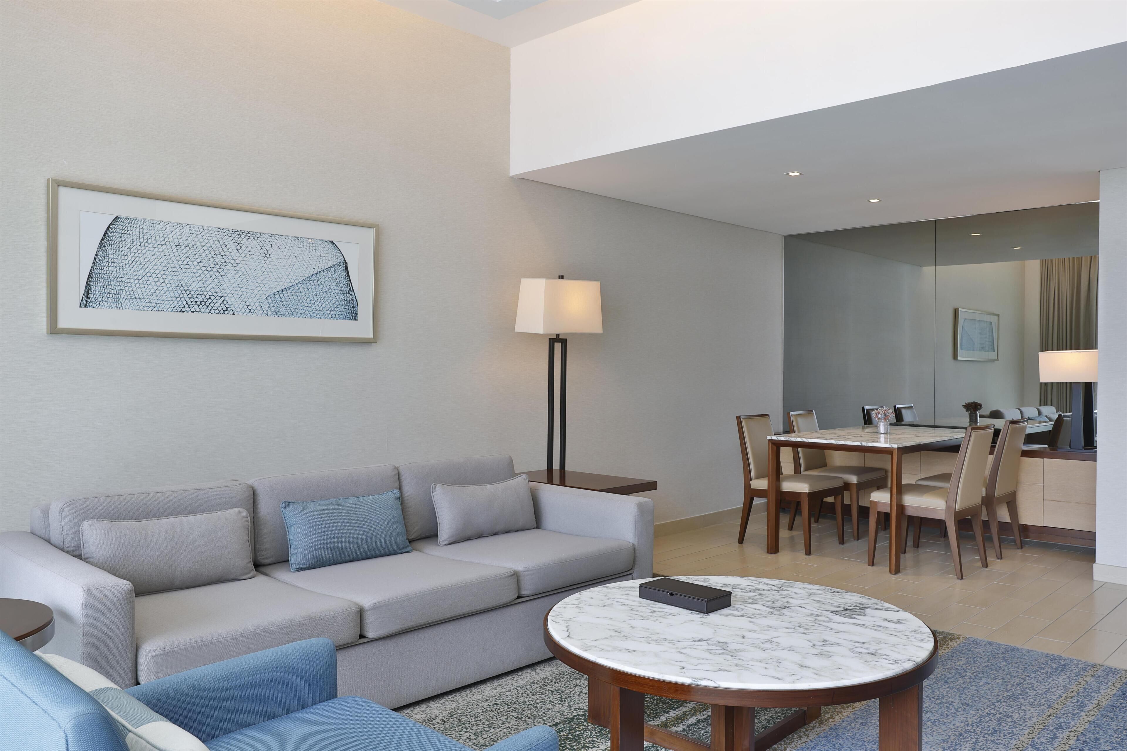 A modern living room with a sofa, a dining table with chairs, a lamp, and a framed artwork on the wall.