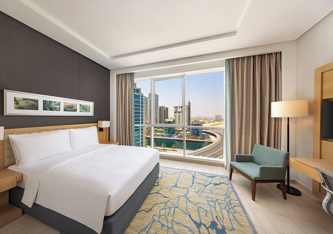 A hotel room with a large bed, a view of the cityscape, a desk, and a chair.
