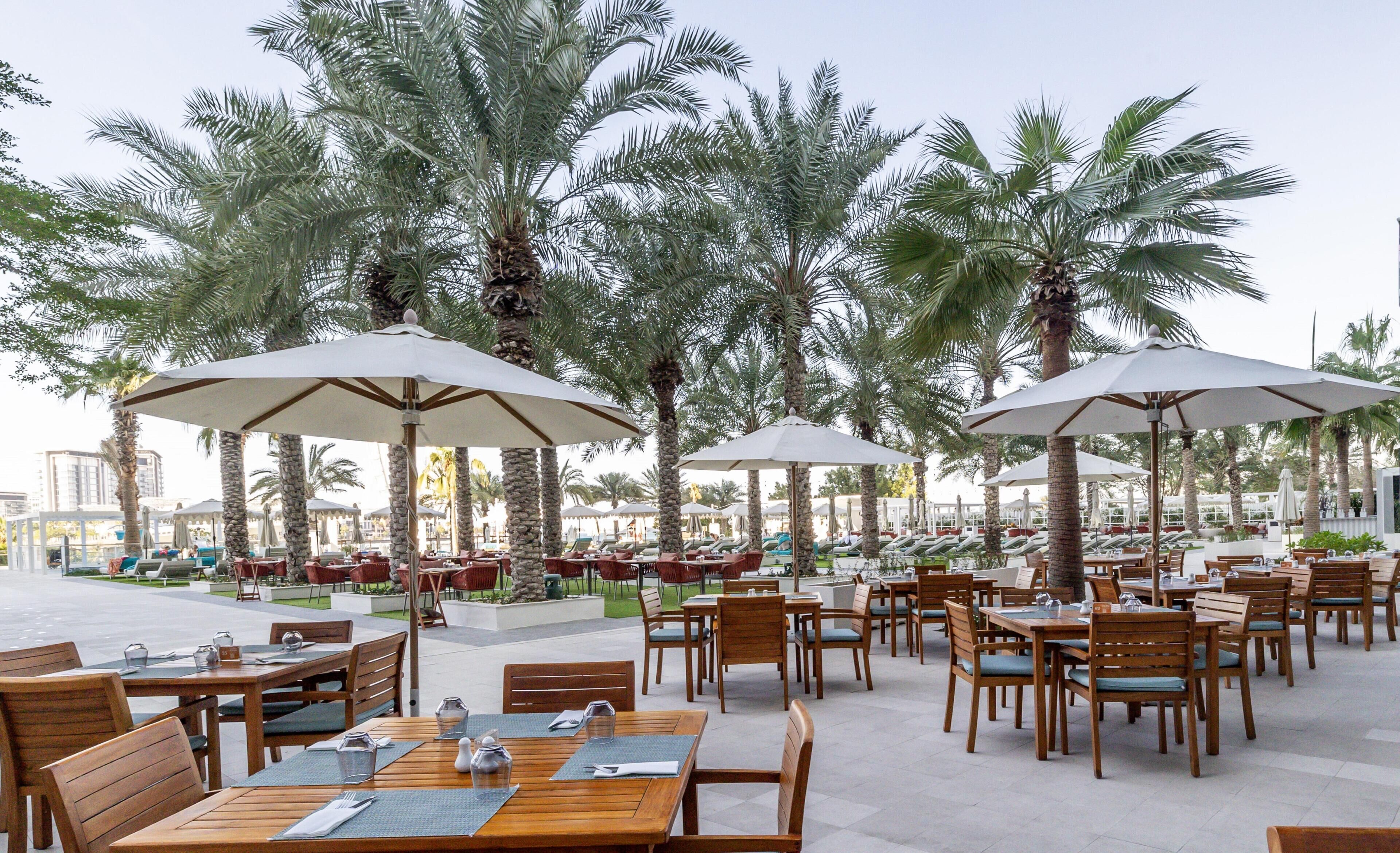 An outdoor dining area with wooden tables and chairs, large umbrellas, and palm trees.