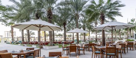 An outdoor dining area with wooden tables and chairs, large umbrellas, and palm trees.