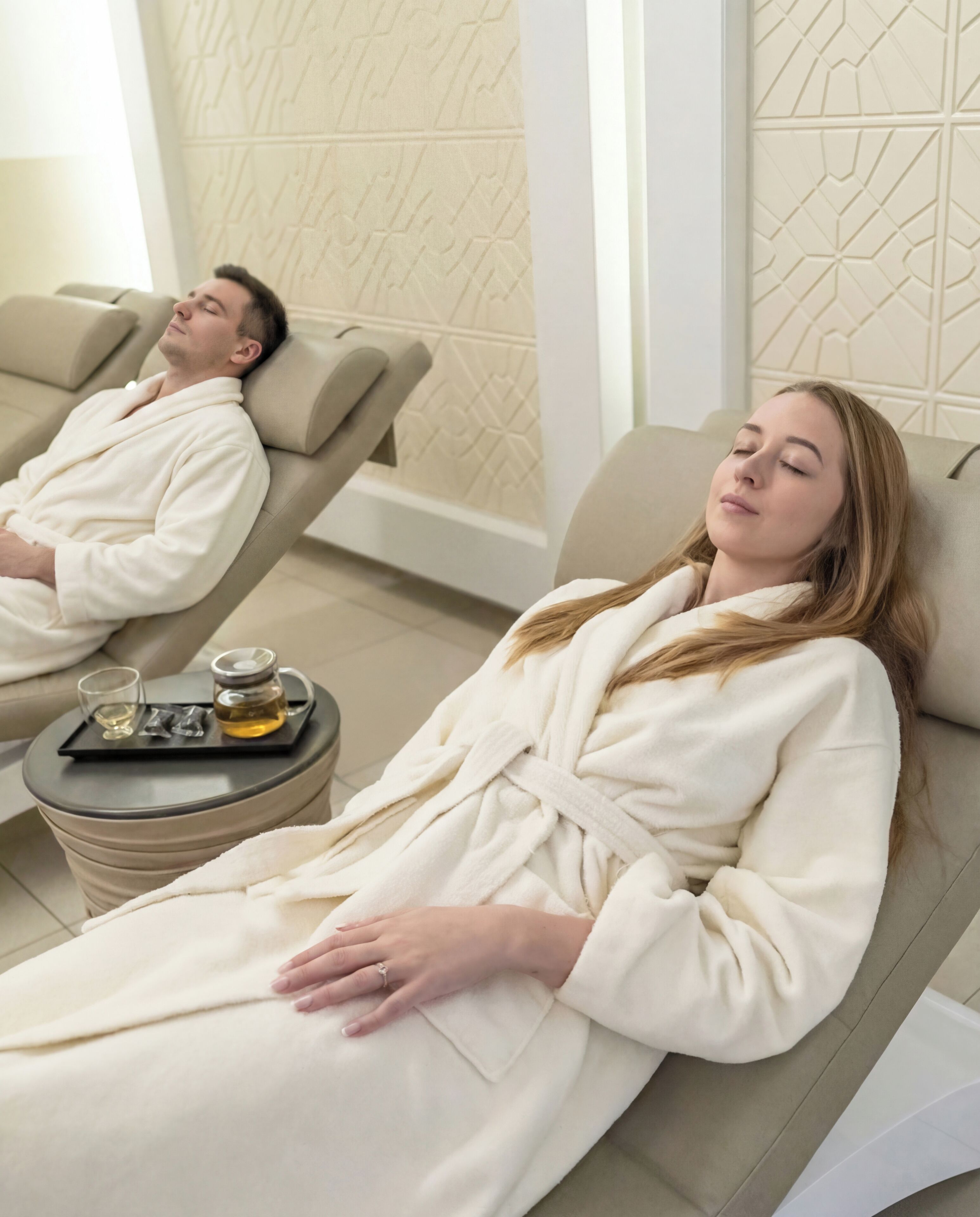 Two individuals in a spa setting, reclining in separate chairs, with a tray of amenities on a small table between them.