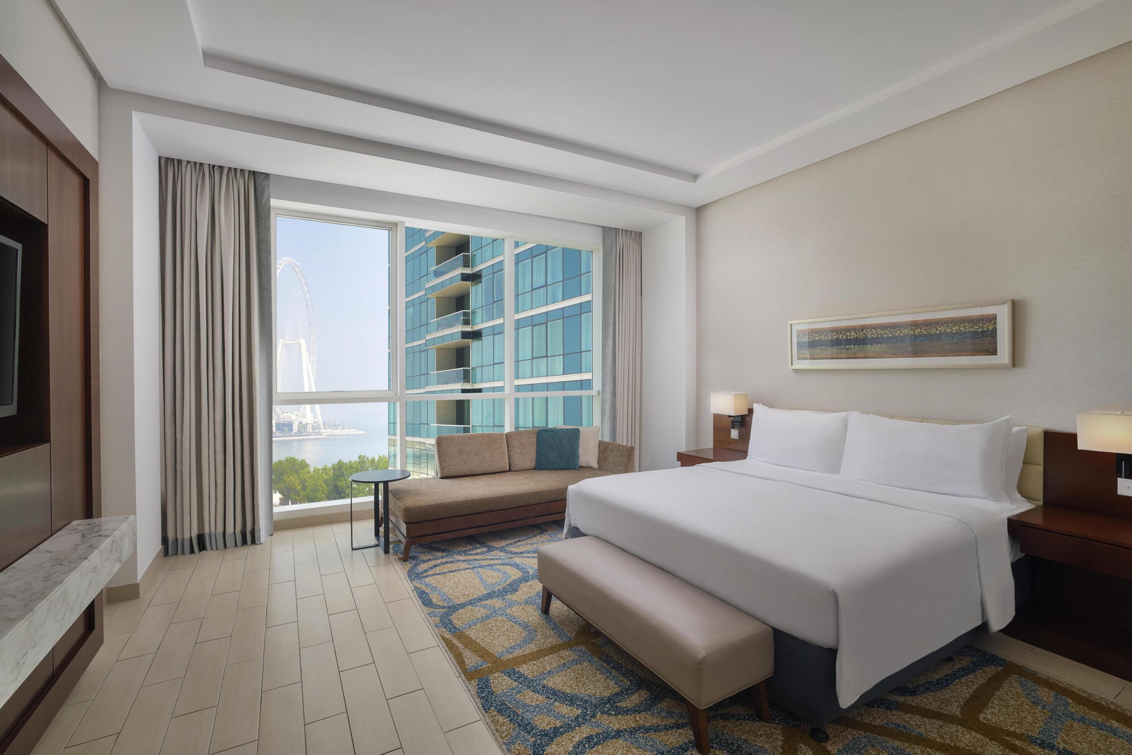 A hotel room with a large bed, a sofa, a small table, a TV, and a view of a building and a bridge.
