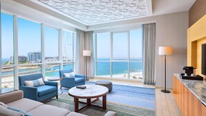 Une chambre d’hôtel moderne dotée de grandes fenêtres, d’un canapé, d’une table basse et offrant une vue sur la mer et le paysage urbain.