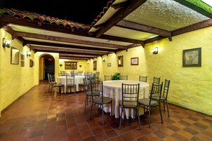 Un espacio exterior para comer con varias mesas y sillas, piso de baldosas y una pared amarilla con cuadros enmarcados.