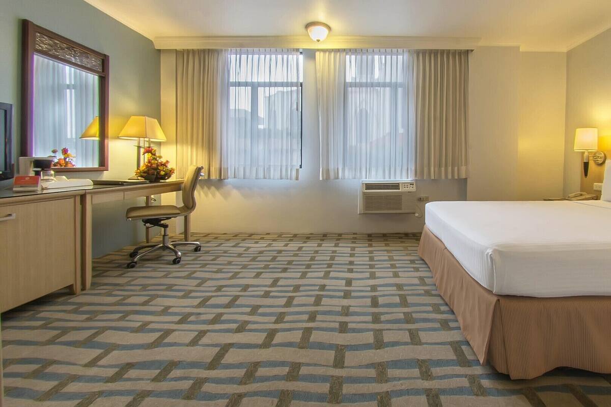 Een hotelkamer met een bed, bureau, stoel, airconditioning en een schilderij aan de muur.