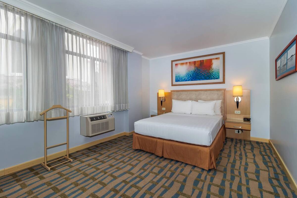 Een hotelkamer met een bed, twee bedlampen, een raam met gordijnen, een airconditioningsysteem en een schilderij aan de muur.