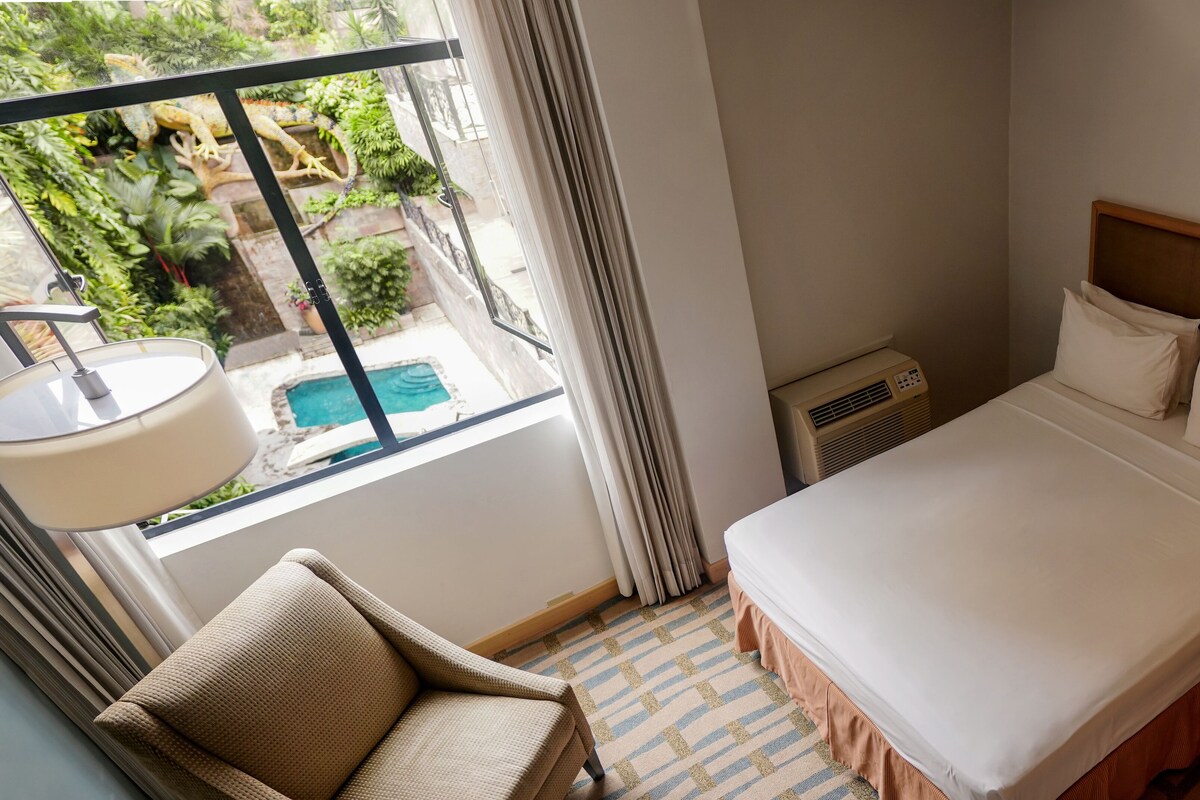 A hotel room with a bed, a chair, a window with a view of a pool and greenery, and an air conditioning unit.