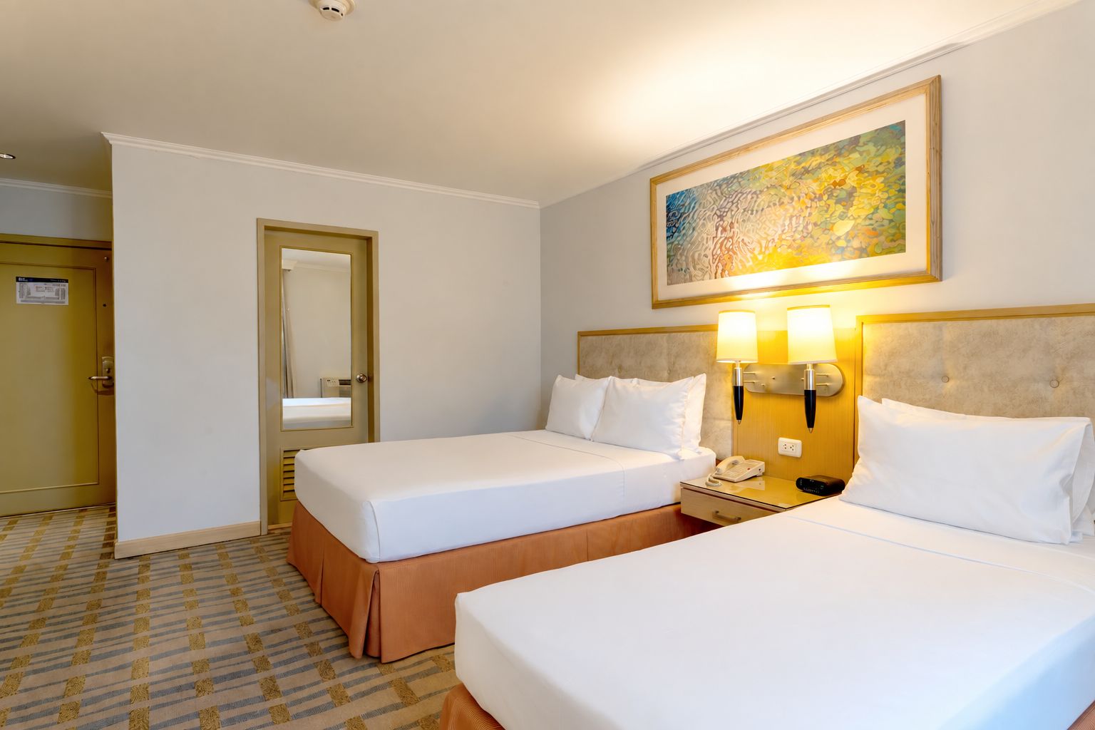 A hotel room with two beds, a bedside table with a lamp, a telephone, and a painting on the wall.