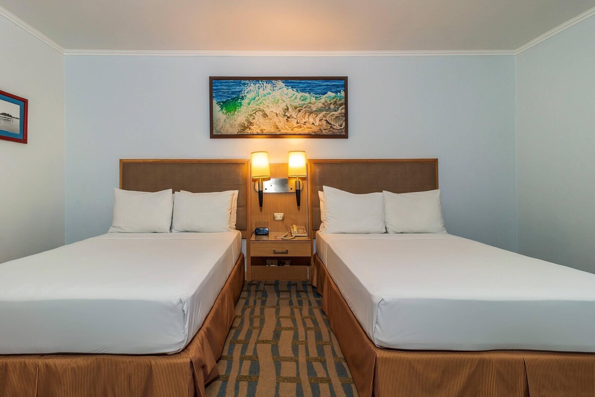Een hotelkamer met twee bedden, een nachtkastje met een lamp, een schilderij van een golf en een ingelijste foto aan de muur.