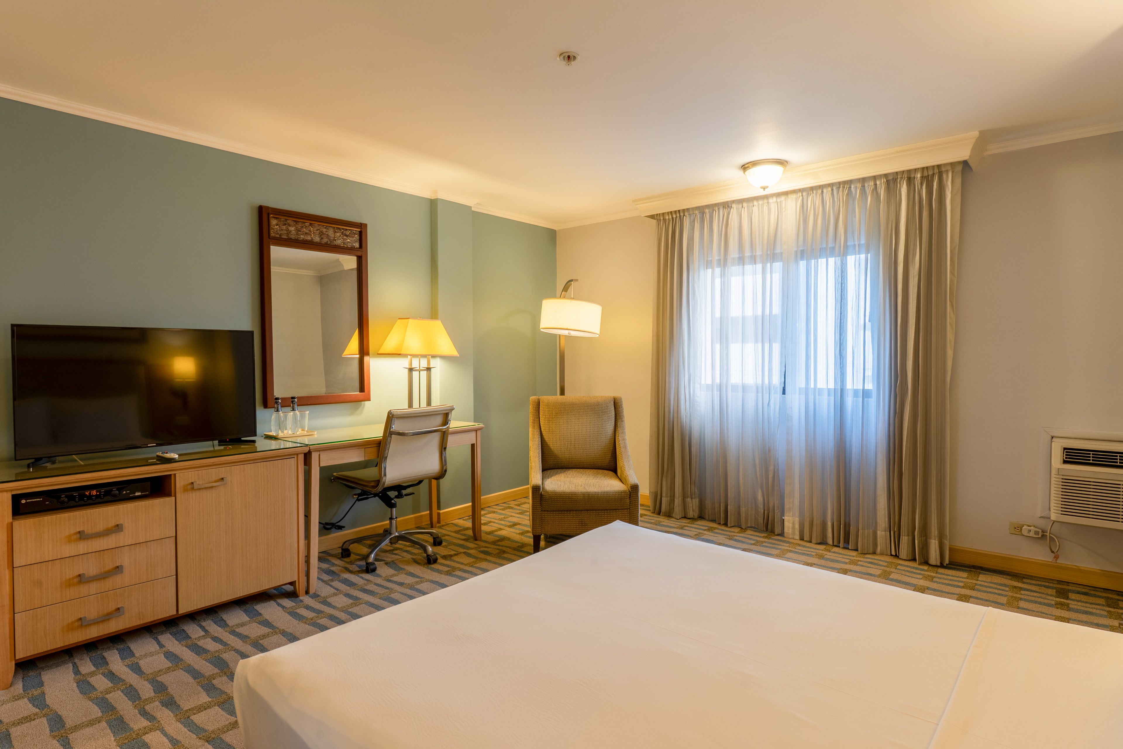 Habitación de hotel con una cama, un escritorio con silla, televisor, espejo, lámpara y ventana con cortinas.