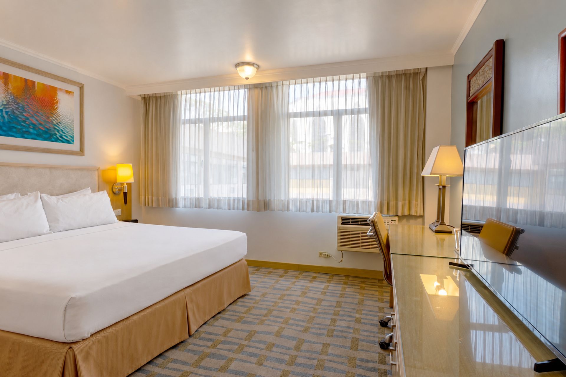 Un cuarto de hotel con una cama grande, dos lámparas de noche, un escritorio con una silla, una ventana con cortinas y un cuadro en la pared.