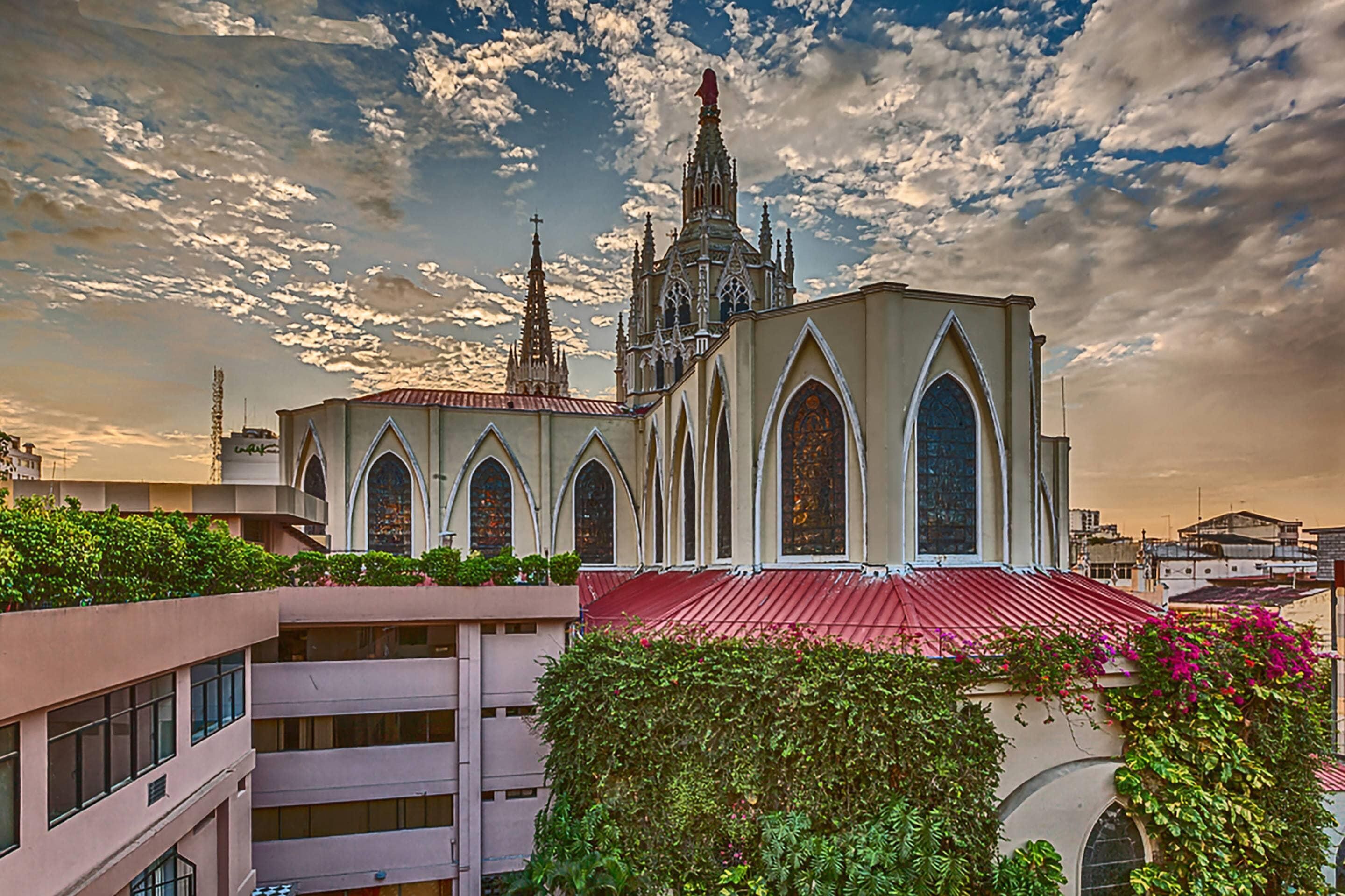 A historic church with a red roof, surrounded by greenery and modern buildings.