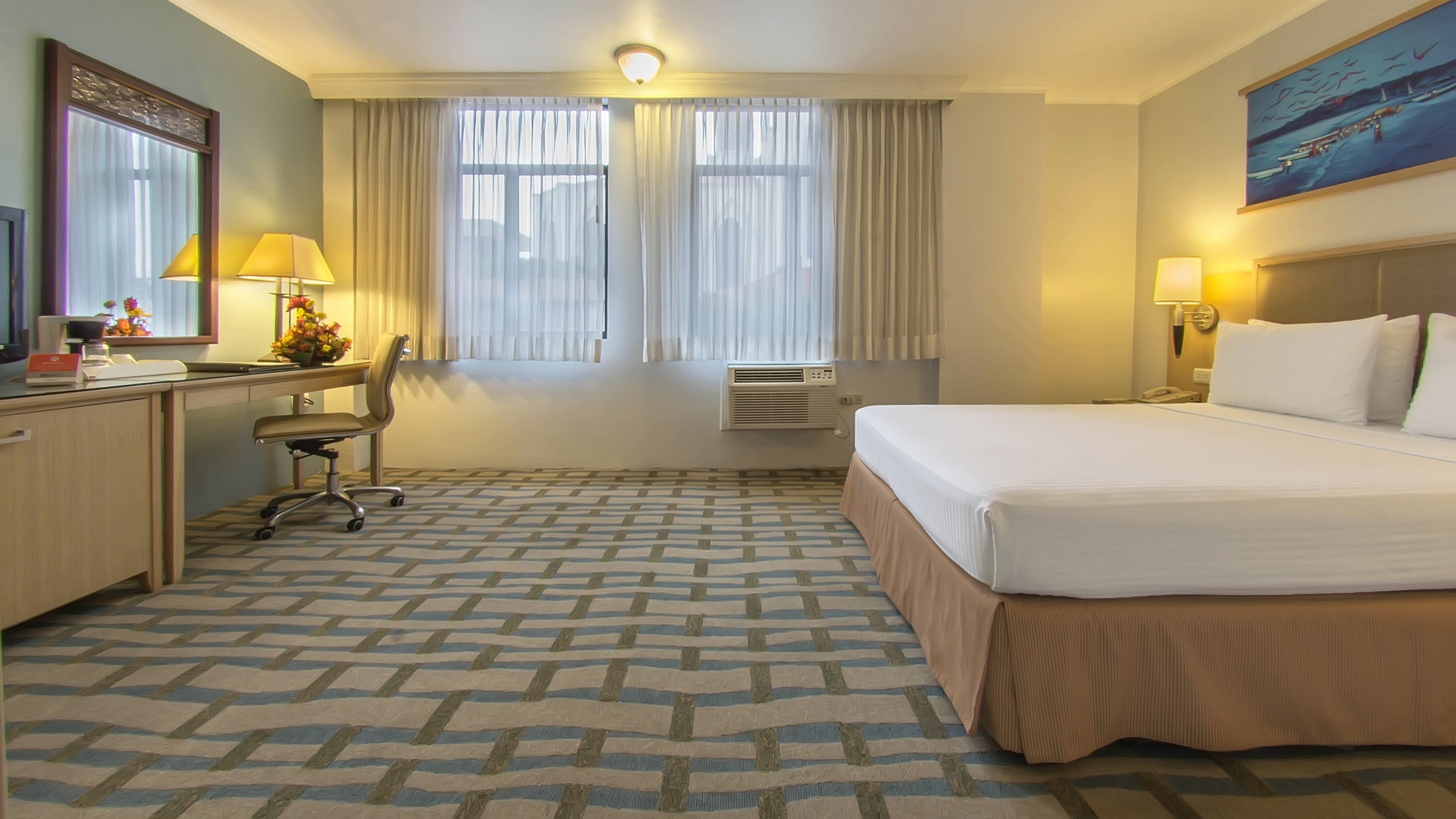Una habitación de hotel con cama, escritorio, silla, aire acondicionado y un cuadro en la pared.