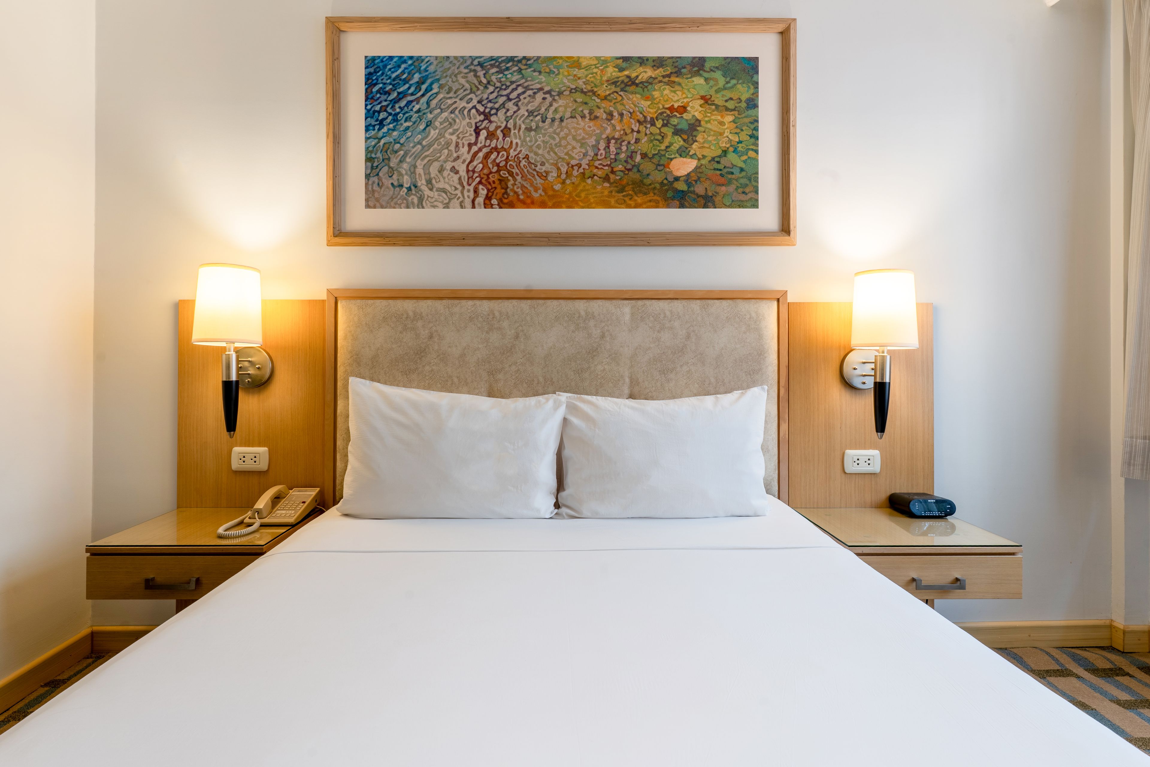 Una habitación de hotel con una cama grande, dos lámparas de noche, un teléfono y un cuadro sobre la cama.