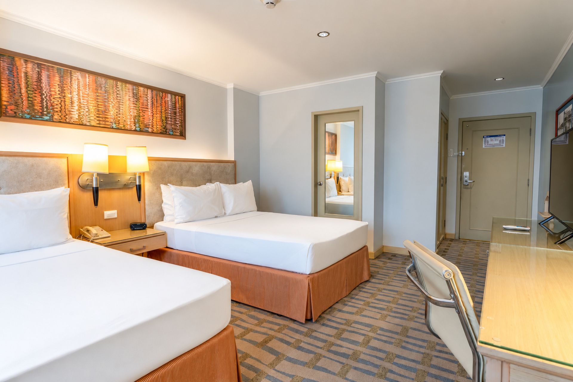 Une chambre d’hôtel avec deux lits, un bureau et une chaise. Il y a un tableau au mur et une porte qui donne sur une autre pièce.
