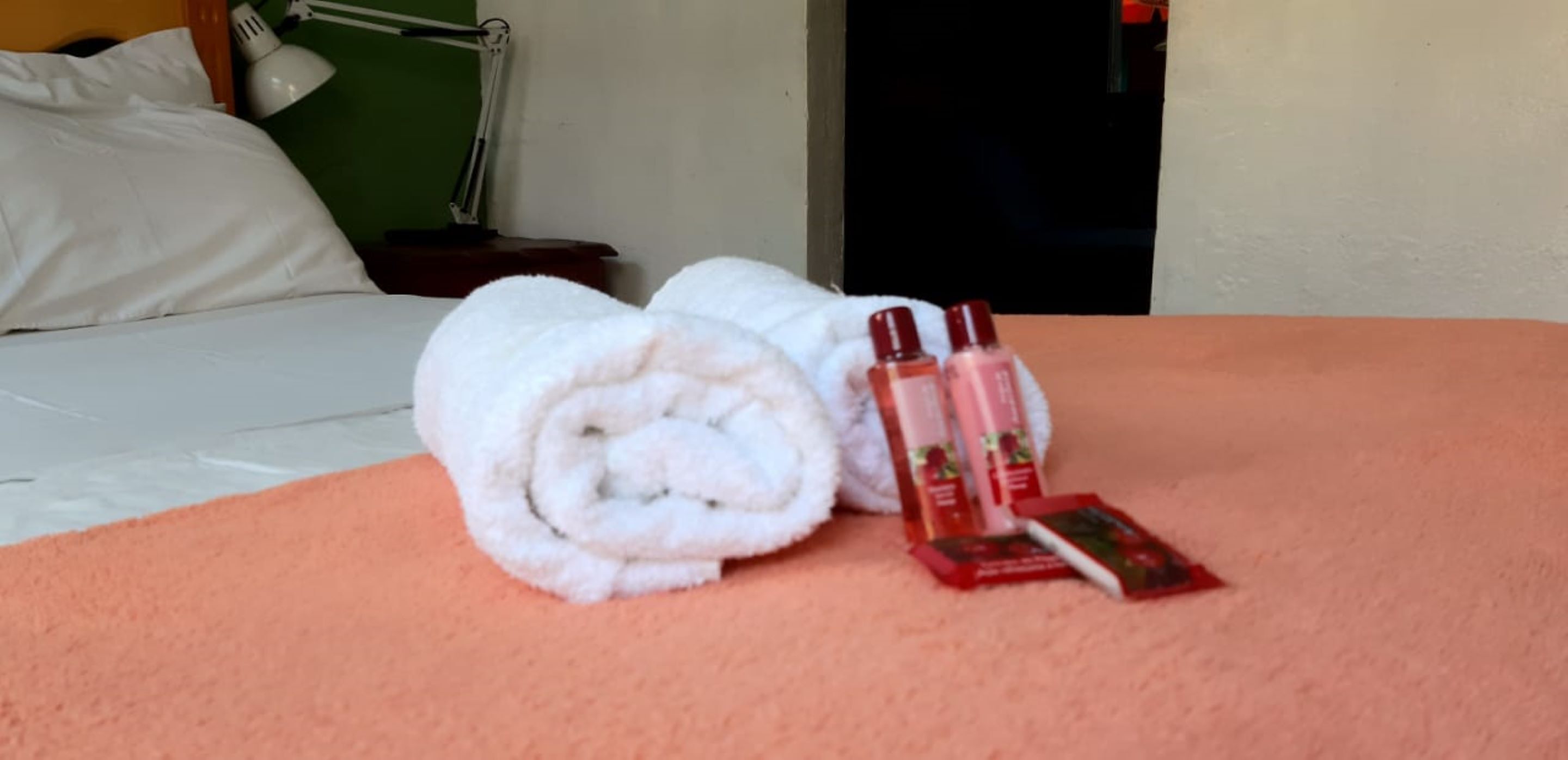 Cama bem arrumada com uma toalha branca enrolada e duas embalagens de esmalte vermelho sobre um cobreleiro laranja.