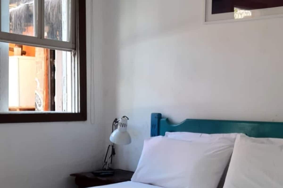 A neatly made bed with white and orange bedding, a bedside lamp, and a window with a view of the outside.